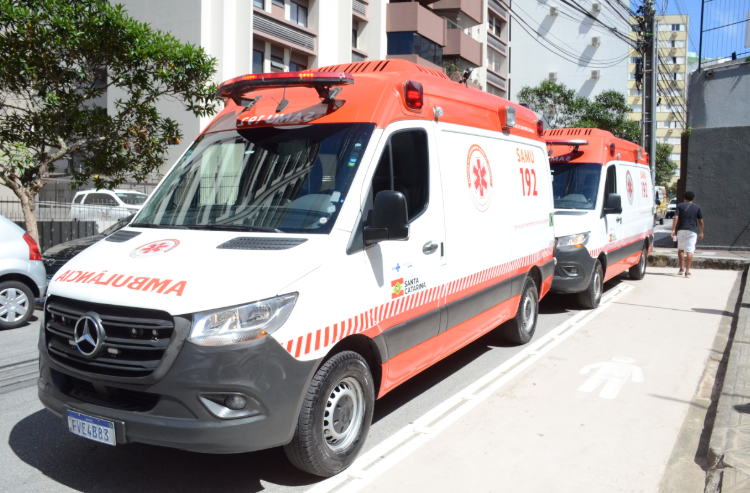 A imagem mostra duas ambulâncias do SAMU (Serviço de Atendimento Móvel de Urgência) estacionadas em fila ao lado de uma calçada. Os veículos são modelos Mercedes-Benz Sprinter, pintados de branco com detalhes em vermelho e a inscrição “SAMU 192” nas laterais, além do logotipo do serviço e a indicação de “Santa Catarina”. Na frente, a palavra “AMBULÂNCIA” aparece invertida para facilitar a leitura em retrovisores. O local parece ser uma área urbana, com prédios residenciais e árvores ao fundo. À direita da imagem, uma pessoa caminha pela calçada, de costas para a câmera.
