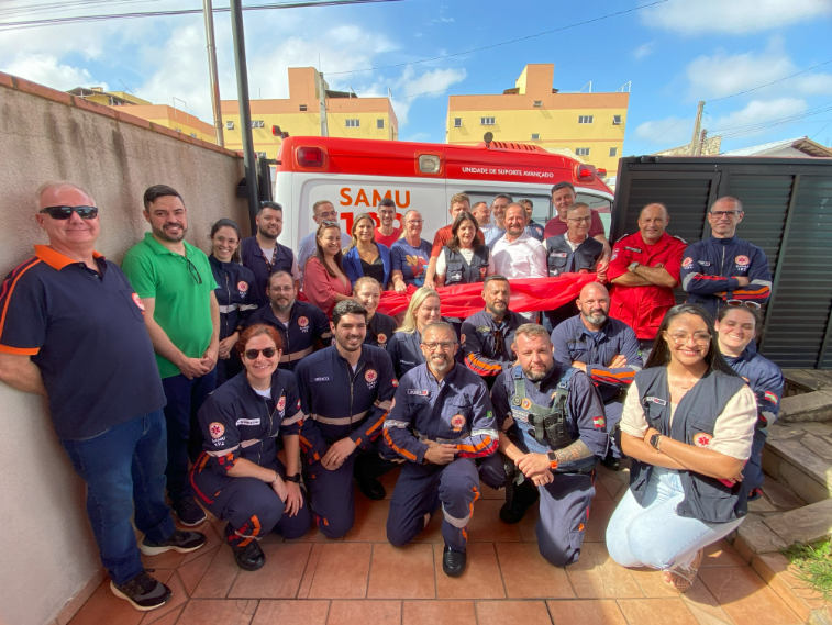 A imagem mostra um grupo de aproximadamente 25 pessoas posando para foto em frente a uma ambulância do SAMU 192 (Serviço de Atendimento Móvel de Urgência), identificada como Unidade de Suporte Avançado. A maioria das pessoas veste uniformes azul-marinho com detalhes em laranja e o emblema do SAMU, indicando que são profissionais de saúde e resgate. Alguns usam roupas casuais e outros vestem camisetas ou jalecos com identificação do serviço. Ao centro, há uma faixa vermelha sendo segurada pelo grupo. O cenário é externo, com céu azul e prédios residenciais ao fundo. O clima da foto transmite espírito de equipe e união.