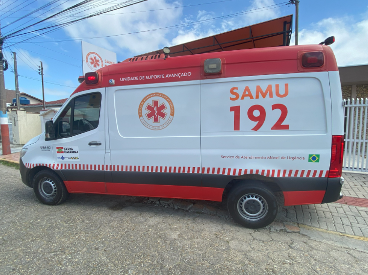 A imagem mostra uma ambulância do SAMU 192 (Serviço de Atendimento Móvel de Urgência) estacionada em via urbana. O veículo é identificado como Unidade de Suporte Avançado (USA-03) de Mafra, Santa Catarina, com pintura branca e faixas vermelhas. Na lateral, há o logotipo do SAMU, a bandeira do Brasil, o número de emergência (192) e menções ao SUS e ao Governo de Santa Catarina. O cenário ao fundo inclui calçada, muro, portão branco e parte de construções residenciais, com céu parcialmente nublado.