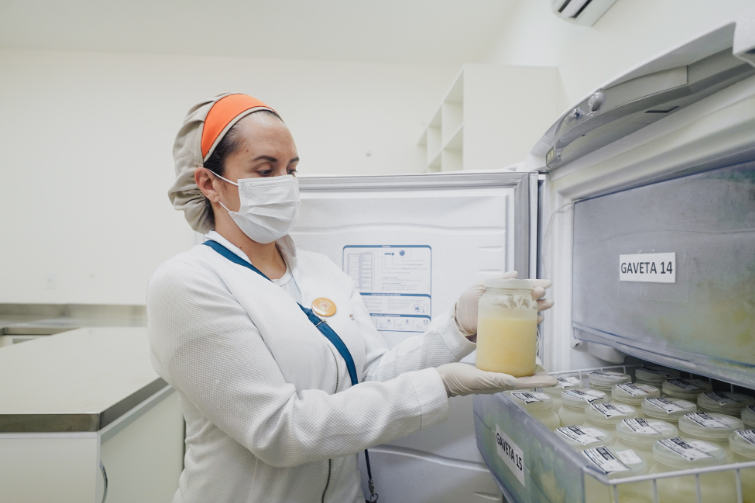A imagem mostra uma profissional de saúde, usando touca, máscara, luvas e jaleco, manuseando um pote de vidro contendo leite humano armazenado. Ela está diante de um freezer aberto, com prateleiras organizadas e rotuladas, visíveis as etiquetas “GAVETA 14” e “GAVETA 15”, contendo vários frascos semelhantes. O ambiente é limpo e iluminado, com aparência de laboratório ou banco de leite humano.
