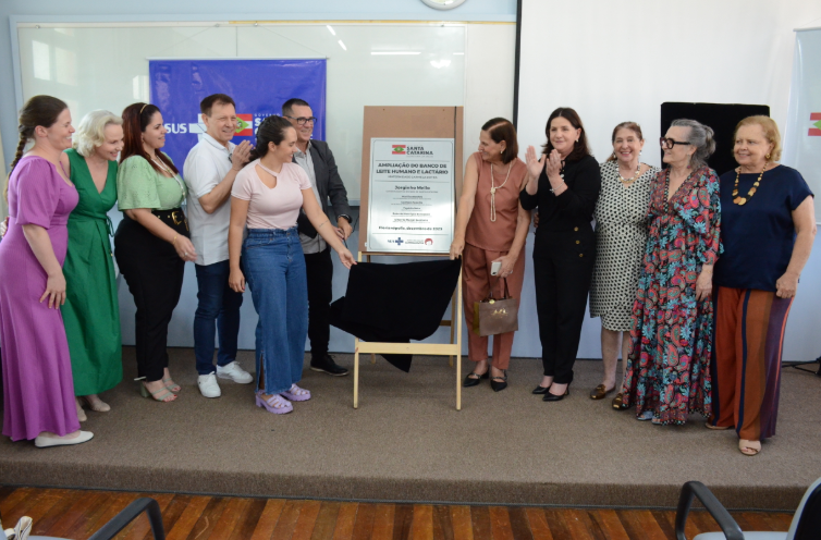 A imagem mostra um grupo de pessoas reunidas em um palco, participando de uma cerimônia de inauguração ou ampliação. No centro, duas mulheres retiram um pano preto para revelar uma placa que menciona a “Ampliação do Banco de Leite Humano e Lactário” em Santa Catarina, com data de dezembro de 2023. Ao fundo, há um banner azul com o logotipo do SUS e do Governo de Santa Catarina. As pessoas ao redor, vestidas de forma variada e elegante, sorriem e aplaudem, demonstrando clima de celebração e reconhecimento.