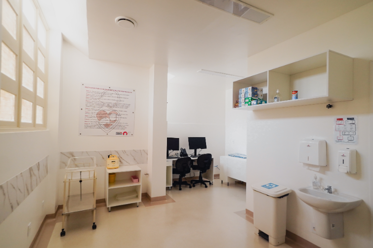 A imagem mostra um ambiente claro e bem organizado, possivelmente uma sala de apoio em um hospital ou banco de leite humano. À esquerda, há uma janela grande que permite entrada de luz natural e, na parede, um cartaz com o título “Dez passos para o sucesso do aleitamento materno”. Abaixo, vê-se um carrinho com estrutura metálica e um móvel pequeno contendo um frasco coletor e outros itens.  Ao centro, em um espaço mais ao fundo, há duas cadeiras giratórias diante de computadores e telefone sobre uma bancada, sugerindo um setor administrativo ou de registro. À direita, sobre a parede, há um armário aberto com caixas de luvas e alguns frascos. Mais abaixo, encontram-se um dispenser de papel toalha, um dispenser de sabonete líquido, uma pia de mãos e uma lixeira com acionamento por pedal, seguindo protocolos de higiene. O ambiente transmite limpeza, funcionalidade e foco em procedimentos padronizados.