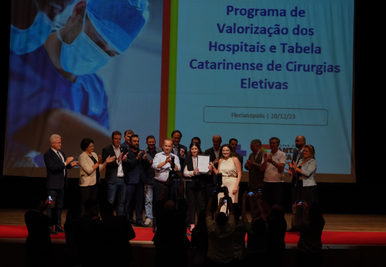 A imagem mostra um grupo de autoridades e representantes reunidos no palco durante um evento. Ao fundo, um telão exibe o título “Programa de Valorização dos Hospitais e Tabela Catarinense de Cirurgias Eletivas”, seguido da localização e data “Florianópolis | 20/12/23”. À esquerda da projeção, há a foto de um profissional de saúde com máscara e touca cirúrgica. No centro do palco, uma mulher segura um documento enquanto é aplaudida, e várias pessoas ao redor também participam da solenidade. Na frente do palco, fotógrafos e participantes registram o momento.