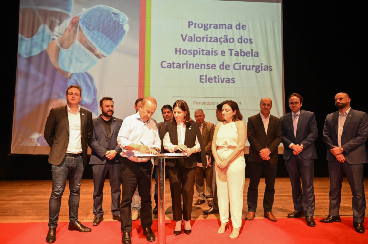 A imagem mostra um evento oficial no palco, com um grupo de autoridades e representantes posicionados em frente a um telão. No fundo, o telão exibe o título “Programa de Valorização dos Hospitais e Tabela Catarinense de Cirurgias Eletivas” e a inscrição “Florianópolis | 20/12/23”. À esquerda da projeção, há a imagem de um profissional de saúde usando máscara e touca cirúrgica.  No centro da cena, um homem assina um documento sobre um púlpito, enquanto uma mulher ao seu lado observa e acompanha o ato. Outras pessoas, vestindo trajes formais, estão posicionadas ao redor, algumas olhando para a assinatura e outras para a plateia. O piso do palco possui um tapete vermelho, destacando a área de assinatura.