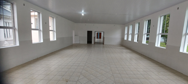 A imagem mostra um salão interno amplo e vazio, com piso de cerâmica clara disposta em padrão diagonal. As paredes são pintadas de branco e possuem janelas grandes nas laterais, permitindo bastante entrada de luz natural. O teto é de forro branco com iluminação simples embutida. Ao fundo, há duas aberturas — uma porta menor e um vão maior — que levam a outro espaço interno. O ambiente parece recém-reformado, limpo e bem iluminado, possivelmente preparado para uso hospitalar ou institucional.