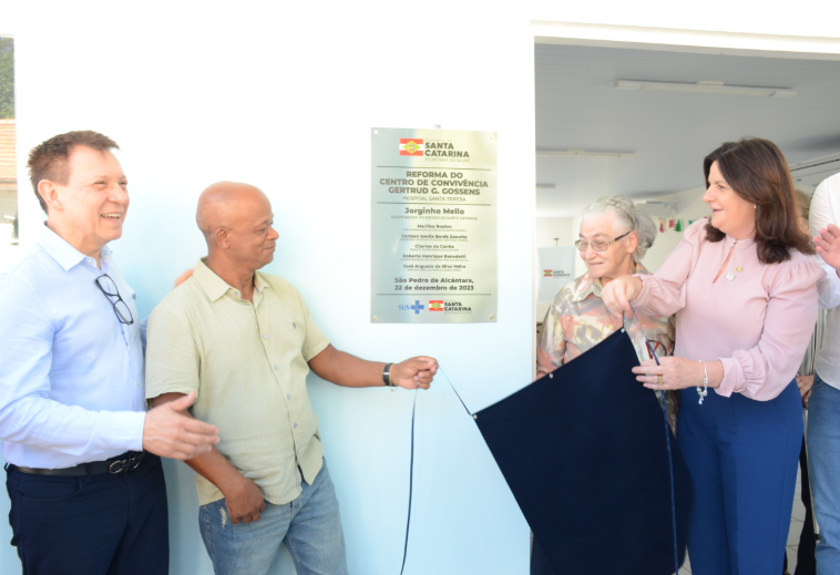 A imagem mostra a cerimônia de inauguração da reforma do Centro de Convivência Gertrud G. Gossens, no Hospital Santa Teresa, em São Pedro de Alcântara.  Quatro pessoas aparecem em destaque, duas de cada lado de uma placa comemorativa afixada na parede. À esquerda, um homem de camisa azul clara e óculos pendurados conversa e sorri, enquanto outro homem de camisa bege segura uma fita. À direita, uma mulher de blusa rosa e calça azul, junto a uma senhora de cabelos grisalhos e óculos, puxa a mesma fita para revelar a placa.  A placa contém informações sobre o evento, incluindo o nome do governador de Santa Catarina, Jorginho Mello, e a data: 22 de dezembro de 2023. O logotipo do SUS e do Governo do Estado de Santa Catarina estão na parte inferior.