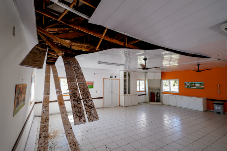 A imagem mostra uma sala ampla e clara com piso de cerâmica branca e paredes pintadas de branco e laranja. O teto está seriamente danificado: parte dele cedeu, revelando a estrutura de madeira e deixando várias placas penduradas e sujas, provavelmente por infiltração ou umidade. No fundo, há armários brancos embutidos, portas e janelas que deixam entrar luz natural. Ventiladores de teto estão instalados, e quadros decoram as paredes, mas o ambiente apresenta um estado de deterioração que indica necessidade urgente de reparos.