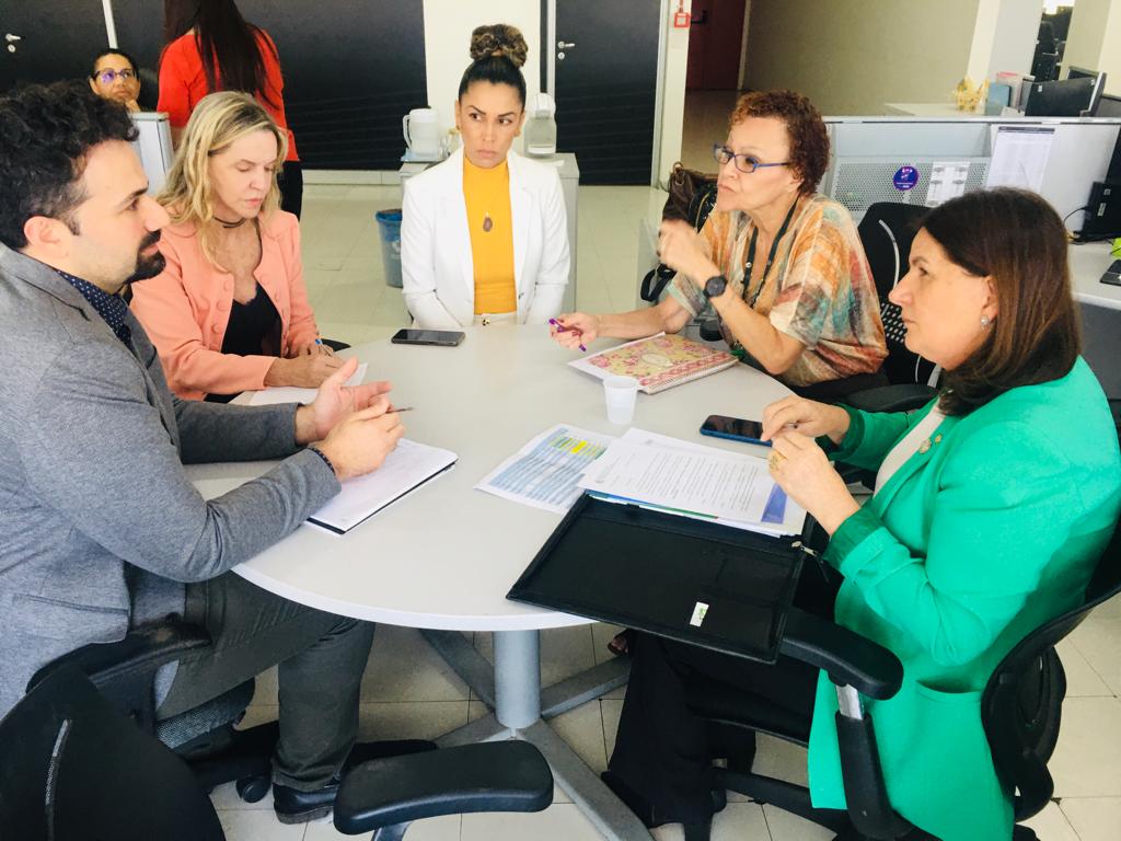 Seis pessoas reunidas ao redor de uma mesa redonda em um escritório. Todos estão concentrados em uma conversa, com documentos, cadernos e celulares sobre a mesa. A mulher com blazer verde parece liderar o diálogo, enquanto os outros escutam e fazem anotações.