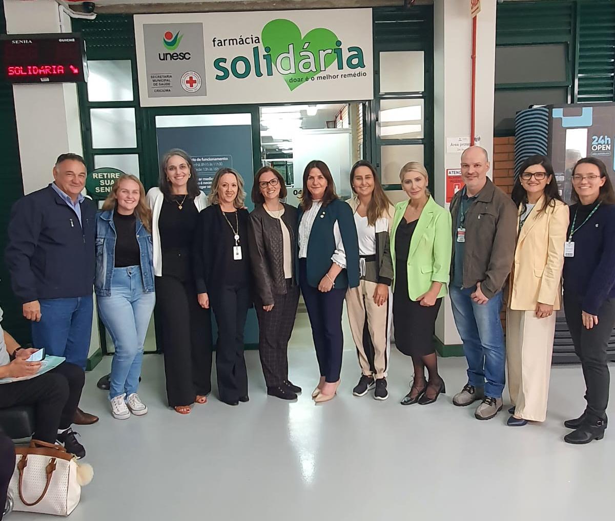 Grupo de pessoas posando para foto em frente à Farmácia Solidária da UNESC, em Criciúma. A placa acima da porta contém os logotipos da UNESC, da Secretaria Municipal de Saúde de Criciúma e da Cruz Vermelha, com o texto 'Farmácia Solidária – doar é o melhor remédio'. As pessoas estão sorrindo, vestidas formalmente, e aparentam estar em visita institucional.