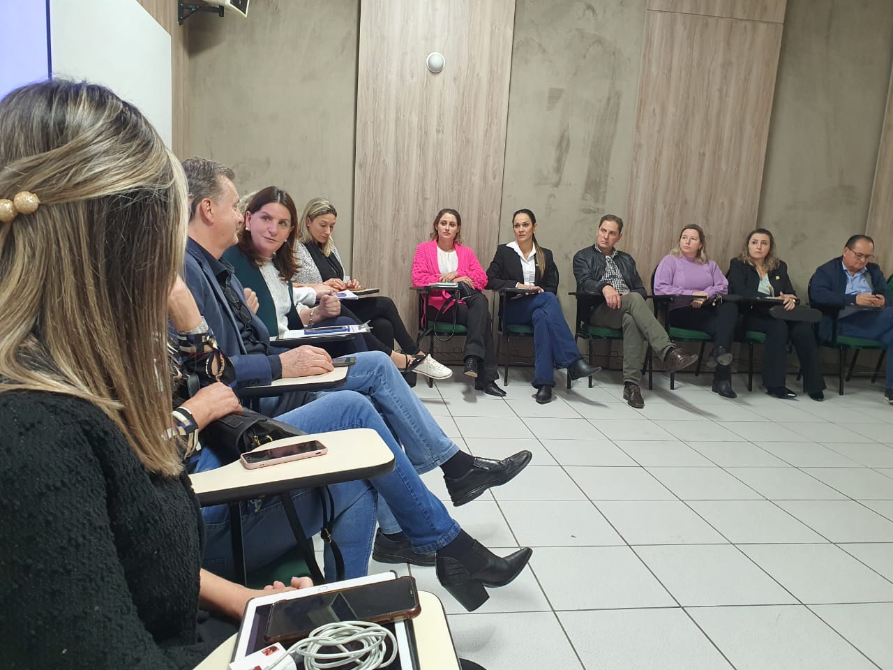 Reunião em sala com formato circular, onde diversas pessoas estão sentadas em cadeiras com pranchetas acopladas, participando de uma conversa. Algumas fazem anotações ou usam celulares. Ambiente com paredes de madeira clara e piso de cerâmica branca. A atmosfera é formal, indicando um encontro institucional ou de planejamento.