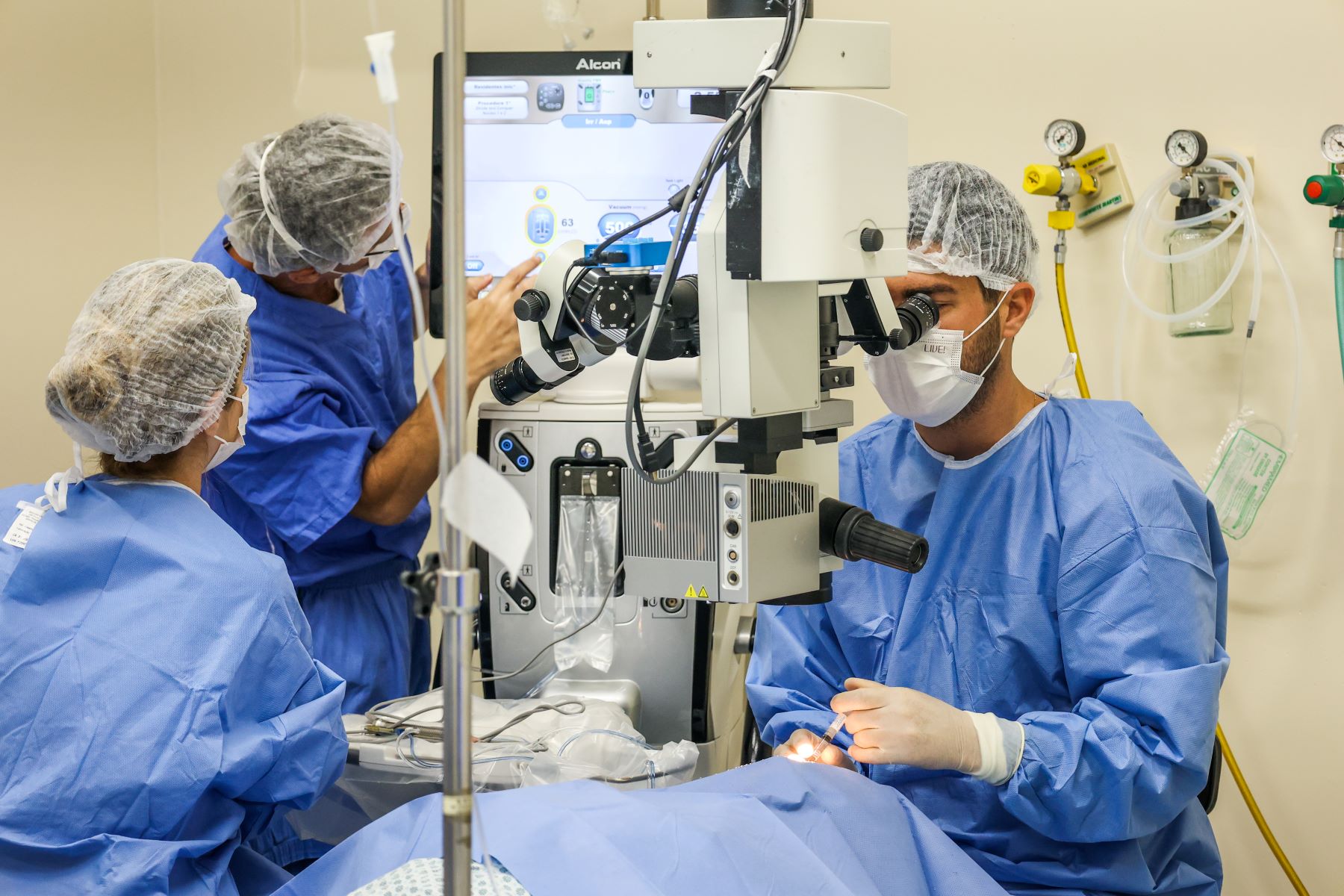 Equipe médica realiza procedimento cirúrgico oftalmológico com auxílio de um moderno microscópio. Três profissionais usam roupas cirúrgicas e toucas, sendo que um opera, outro assiste ao monitor e uma terceira acompanha ao lado. O ambiente é esterilizado, com equipamentos hospitalares visíveis ao fundo.