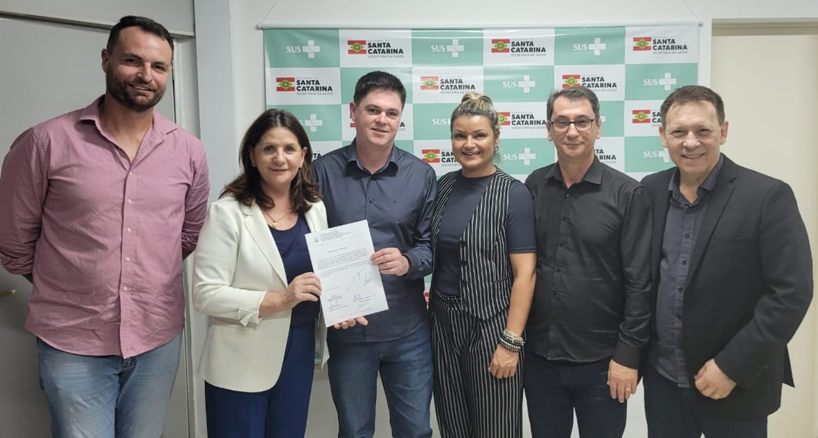 Seis pessoas posando para foto em frente a um painel com a logomarca do SUS e do Governo de Santa Catarina, enquanto duas delas seguram um documento oficial assinado.