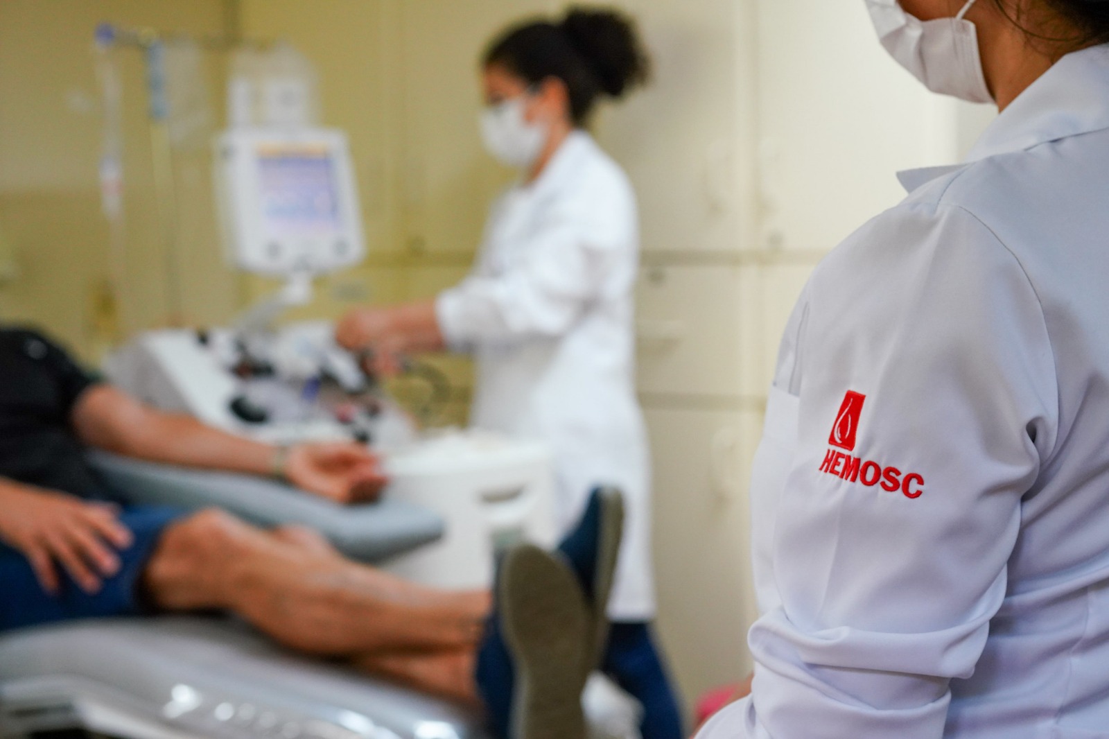Pessoa deitada em cadeira de doação de sangue, com braço estendido ligado a equipamento médico. Ao fundo, profissional de saúde com máscara e jaleco branco ajusta o aparelho. Em primeiro plano, aparece o ombro de outra profissional com jaleco branco, bordado com o logotipo e o nome "HEMOSC" em vermelho.