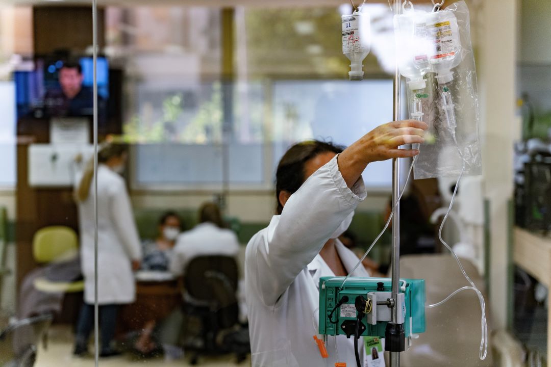 Profissional de enfermagem manuseia frascos de soro em um suporte de infusão em ambiente hospitalar; ao fundo, outros profissionais atendem pacientes em um ambiente clínico.