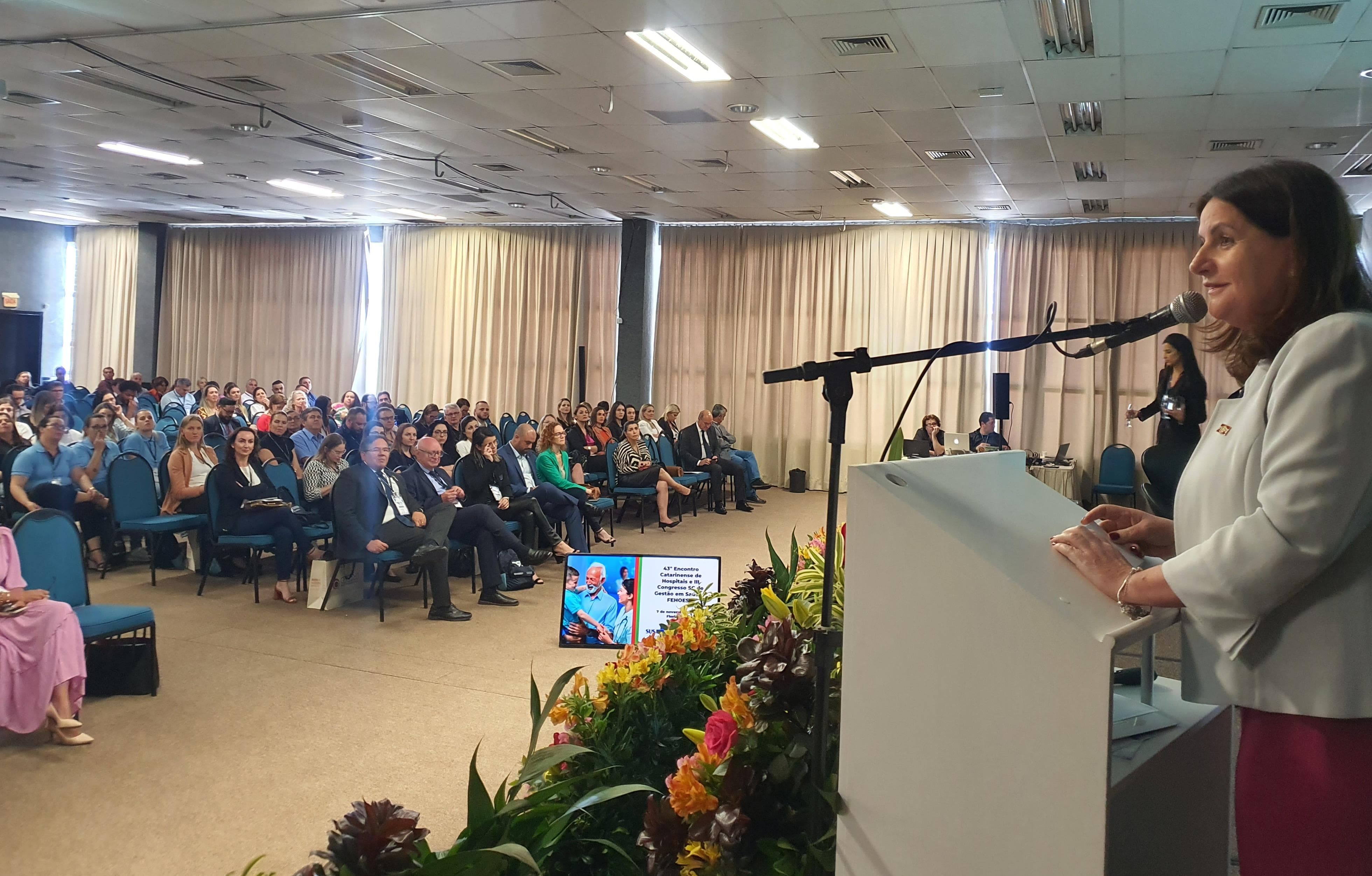 A imagem mostra um evento realizado em um auditório, com um grande número de pessoas sentadas assistindo a uma palestra.  No primeiro plano, à direita, uma mulher está falando ao microfone em um púlpito branco, com arranjos de flores coloridas à frente. Ela veste um blazer claro e parece ser a palestrante principal do momento.  O público, composto por homens e mulheres, está sentado em cadeiras azuis e parece atento. Ao fundo, há uma equipe técnica operando equipamentos de som e projeção. As cortinas claras fecham a iluminação externa, criando um ambiente focado no palco.  No canto inferior, há um monitor exibindo a programação ou informações visuais para os palestrantes.