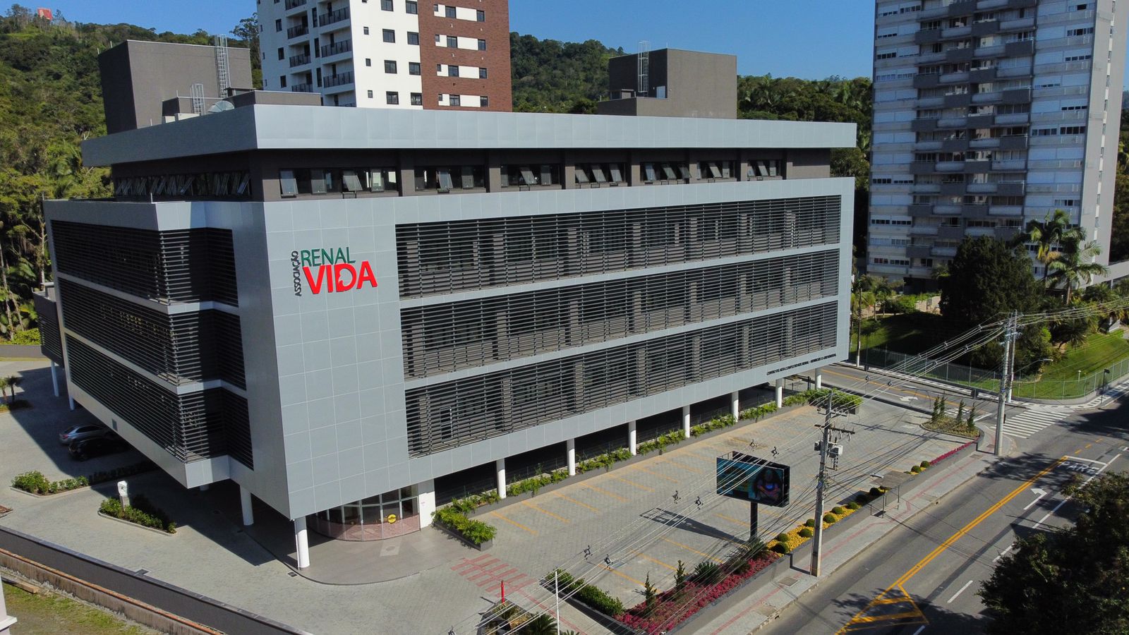 Vista externa de um prédio moderno da "Associação Renal Vida", com fachada cinza e janelas com persianas horizontais. O nome da instituição está em letras grandes na lateral, com as palavras “RENAL” em verde e “VIDA” em vermelho. O edifício está localizado em uma área urbana arborizada, com ruas asfaltadas e outros prédios residenciais ao redor. Há um pequeno estacionamento em frente e uma tela digital na calçada.
