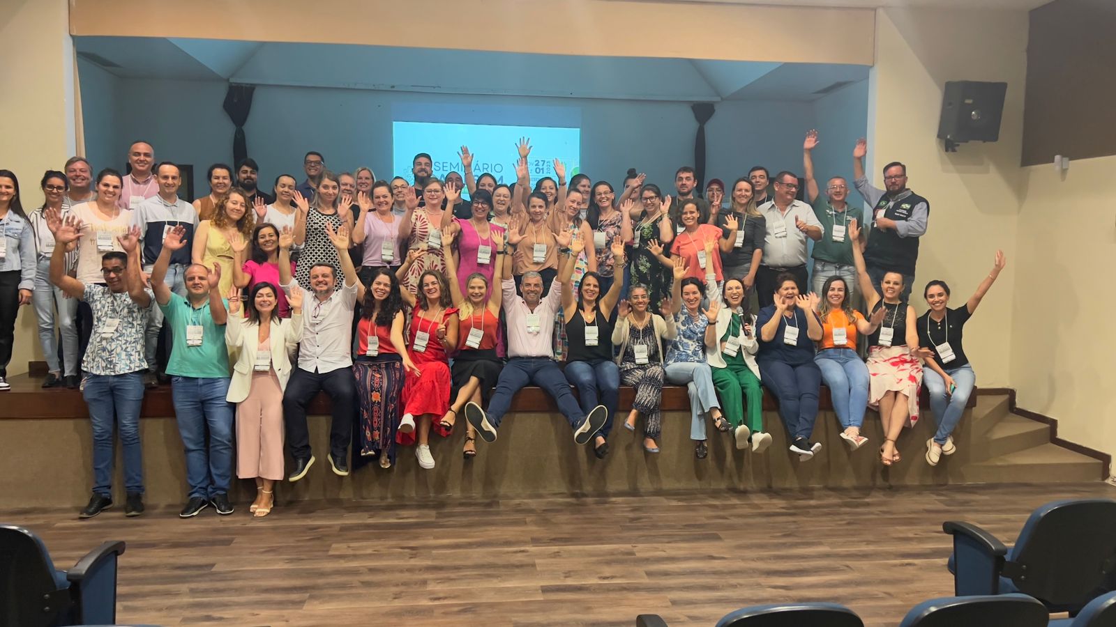 A imagem mostra um grande grupo de pessoas reunidas em um auditório, posando para uma foto coletiva. Todos estão sorrindo e com os braços levantados, transmitindo energia e entusiasmo.  As pessoas estão organizadas em dois níveis: uma parte sentada ou semi-sentada na borda do palco e outra parte em pé atrás. No fundo, há um telão com a projeção que indica tratar-se de um “Seminário” e possivelmente a data “27”. O ambiente é bem iluminado, com piso de madeira e cadeiras azuis visíveis na frente da imagem.  O clima é de confraternização, união e celebração, provavelmente relacionado a um evento de capacitação, reunião de trabalho ou encontro de rede profissional.