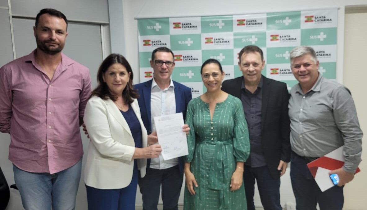 Seis pessoas posando para foto em frente a um painel do SUS e Governo de Santa Catarina, enquanto duas delas seguram um documento oficial assinado.