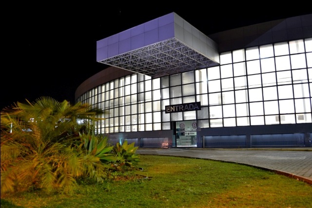 Vista noturna da fachada envidraçada do hospital com letreiro "ENTRADA" acima da porta principal. Estrutura moderna com iluminação interna intensa e jardim com plantas no canto esquerdo.
