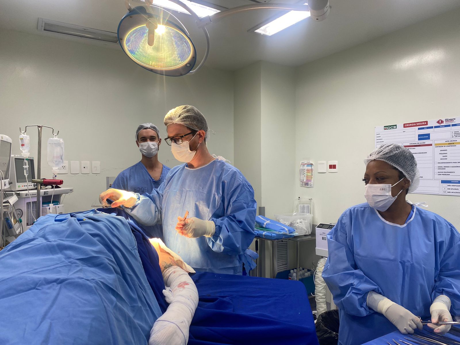 Equipe médica realiza procedimento cirúrgico em centro cirúrgico hospitalar. Profissionais utilizam roupas estéreis, máscaras e toucas, enquanto operam sob iluminação cirúrgica intensa. O ambiente é limpo e equipado com monitores, instrumentos cirúrgicos e materiais hospitalares, refletindo um procedimento técnico em andamento.