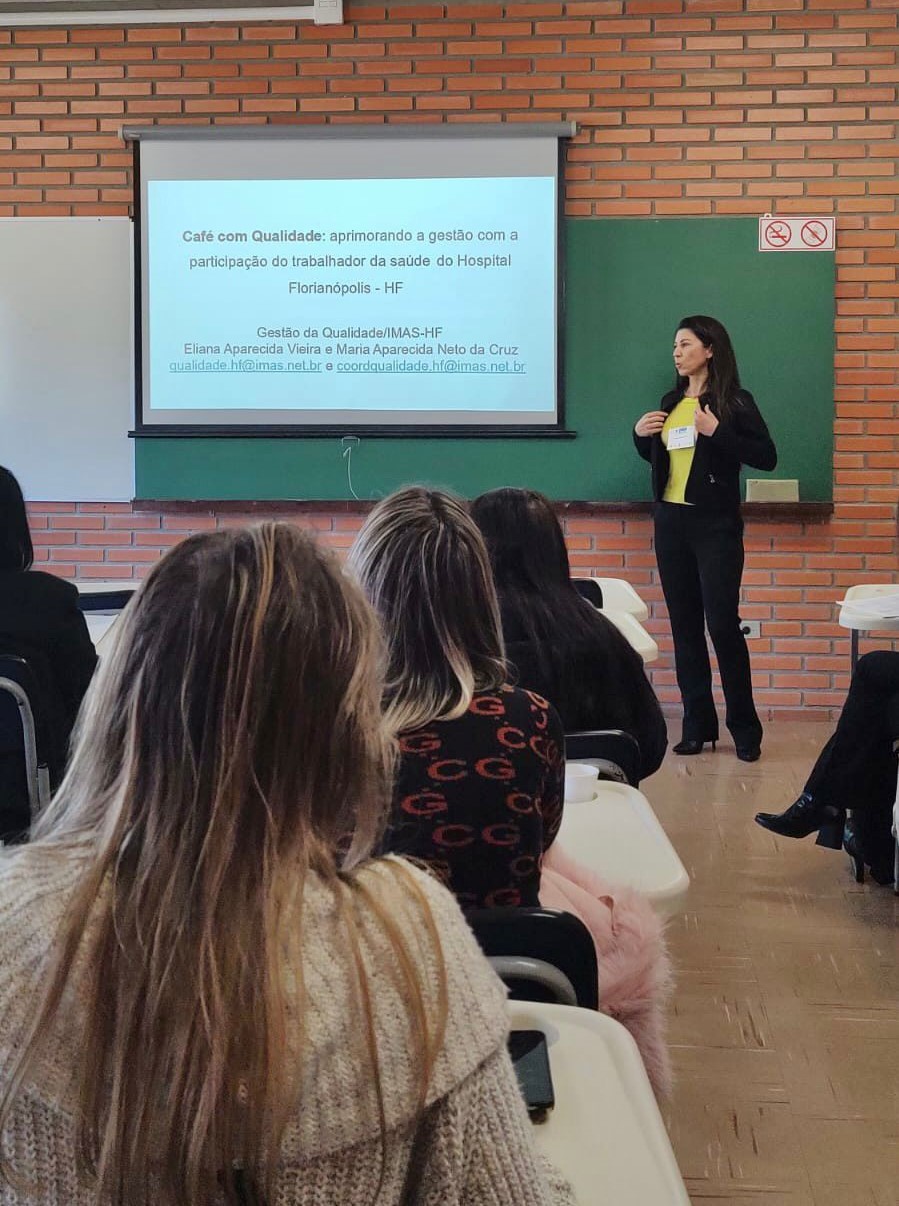 Mulher em pé ao lado de projetor apresentando o tema "Café com Qualidade", sobre a gestão com participação dos trabalhadores da saúde no Hospital Florianópolis. O público está sentado em carteiras escolares, em uma sala com paredes de tijolos à vista.