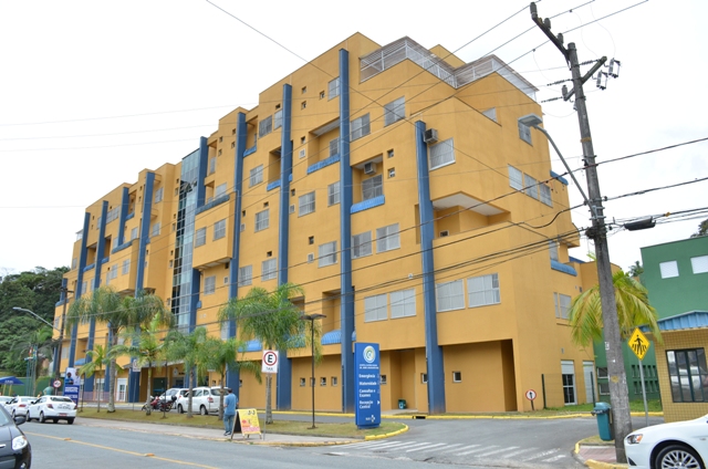 Fachada de um edifício hospitalar de cinco andares com pintura em tons de amarelo e detalhes em azul. Em frente ao prédio, há palmeiras, veículos estacionados e uma placa de identificação do hospital. Fios de energia e postes são visíveis no primeiro plano.