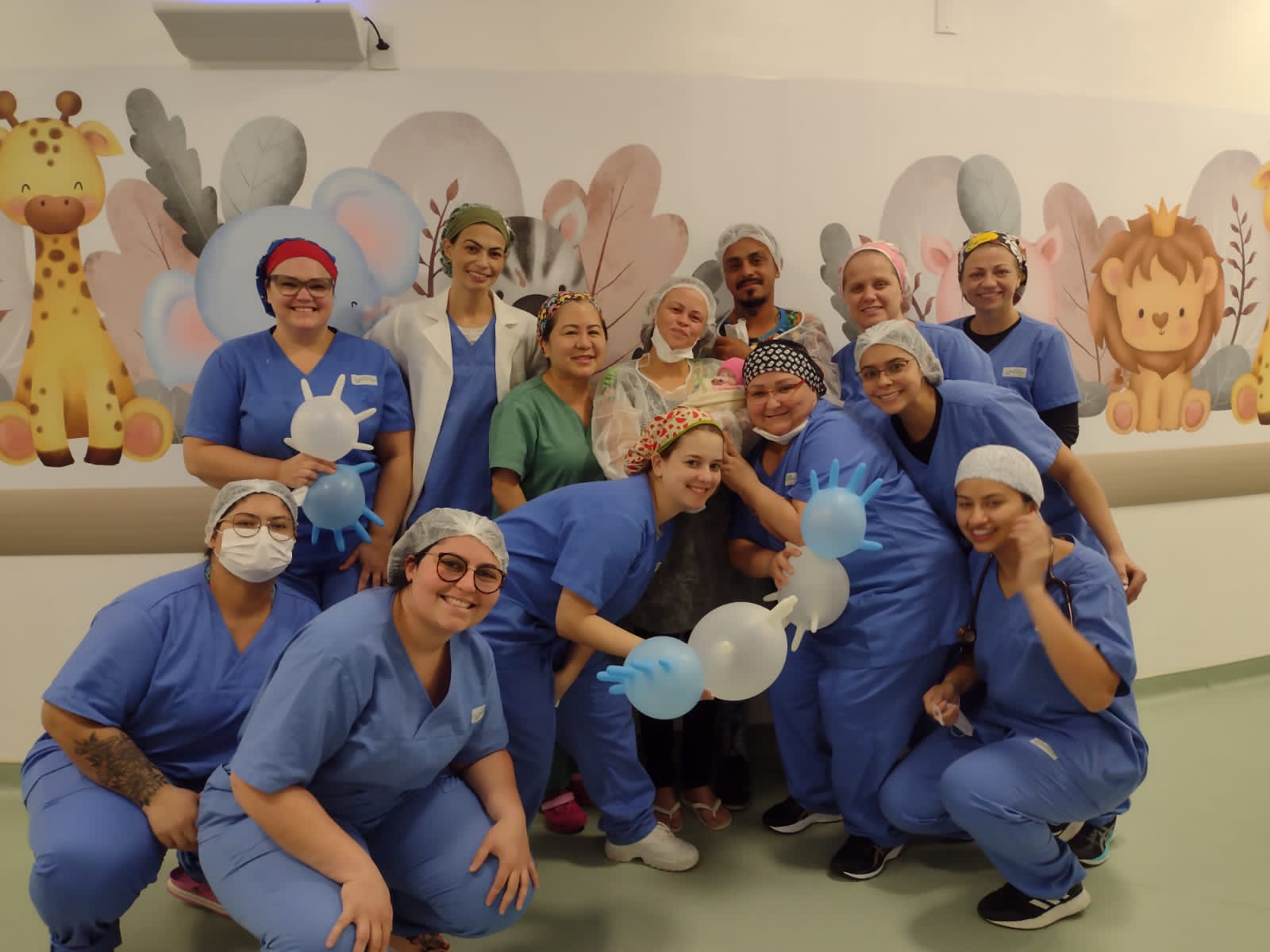 Grupo de profissionais da saúde posa sorrindo em um corredor hospitalar infantil, com parede decorada por ilustrações coloridas de animais como girafa, elefante e leão. As pessoas usam uniformes cirúrgicos azuis e toucas estampadas, e algumas estão com jalecos brancos. No centro da imagem, uma mulher segura uma bebê, que é o foco da homenagem. Balões em formato de luvas médicas estão sendo usados como decoração. O clima é alegre e acolhedor, sugerindo uma celebração ou despedida especial no ambiente hospitalar.