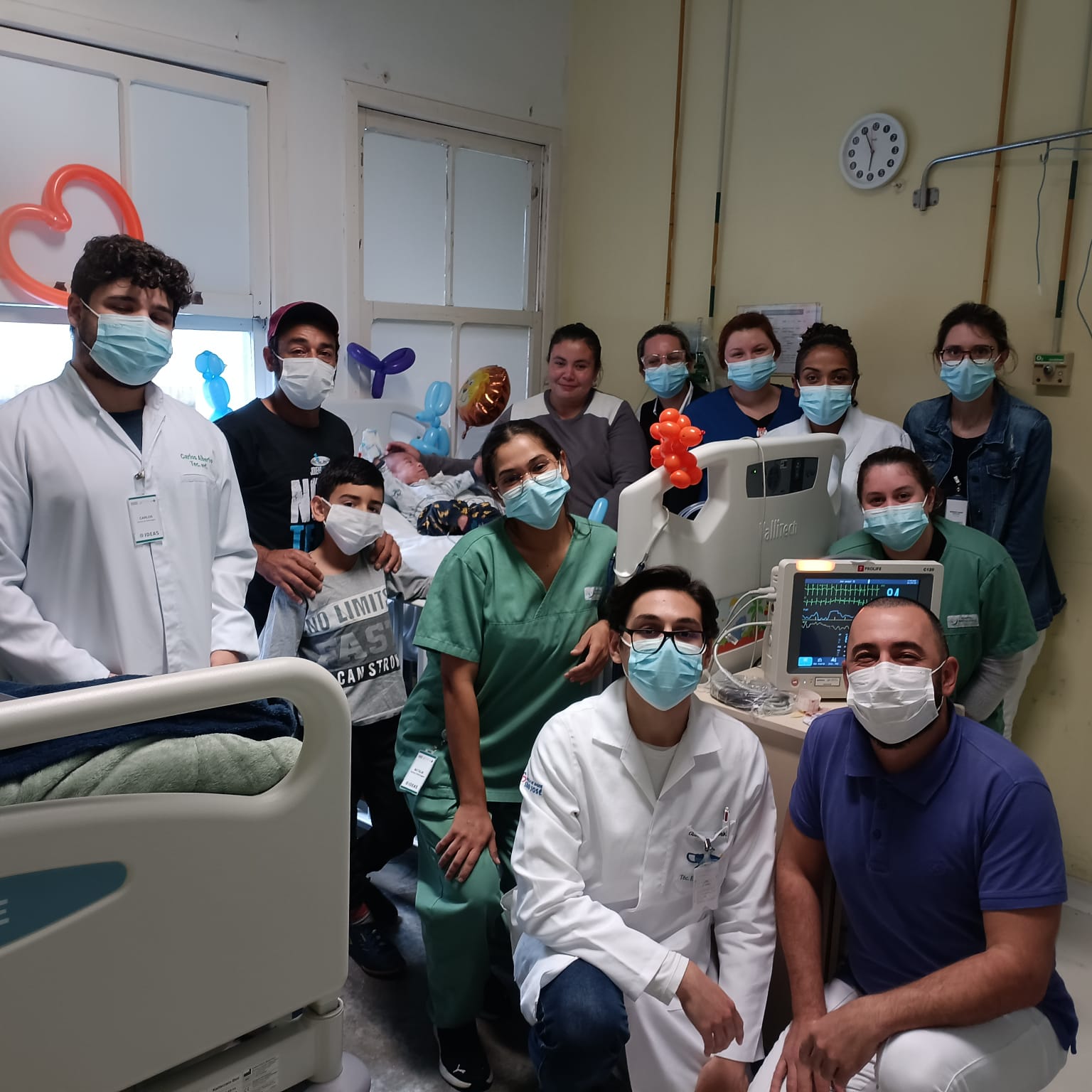 Grupo composto por profissionais da saúde, familiares e uma criança paciente reunidos ao redor de um leito hospitalar pediátrico. Todos estão usando máscaras e demonstram alegria. O quarto está decorado com balões coloridos em forma de animais, criando um ambiente acolhedor e festivo.