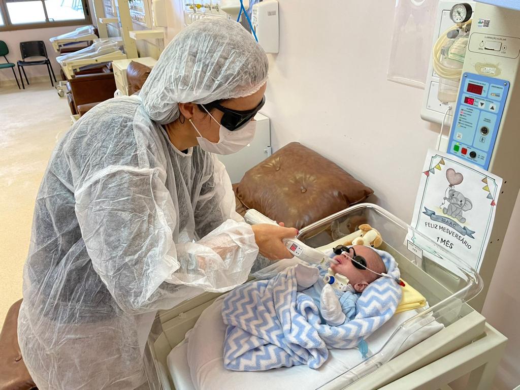 Profissional de saúde, vestindo touca, máscara, óculos de proteção e avental descartável, aplica um tratamento a laser em um bebê recém-nascido deitado em berço hospitalar. O bebê, também com óculos de proteção, está coberto com manta azul e branca. Ao lado, há um cartaz decorado que diz “Feliz Mesversário – 1 mês”.