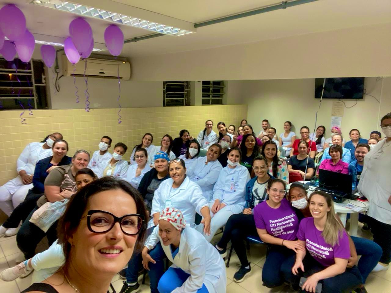 Sala cheia de profissionais de saúde reunidos, muitos usando jalecos brancos e alguns com camisetas roxas com a frase “Prematuridade & UTI Neonatal – Realidade e desafios da ilha”. A pessoa na frente à esquerda tira uma selfie sorrindo, enquanto ao fundo todos olham para a câmera. O teto está decorado com balões roxos pendurados.