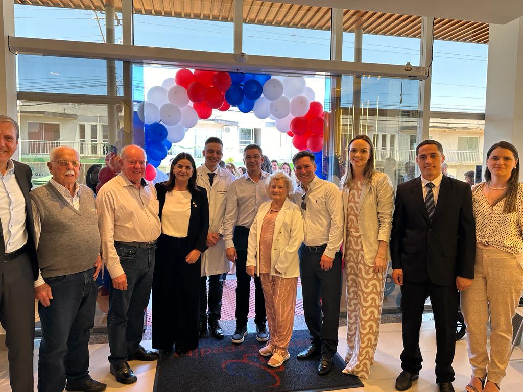 Grupo de pessoas posando para foto na entrada de um prédio decorado com arco de balões nas cores azul, branco e vermelho. Entre eles, há homens de terno, pessoas de roupa social e profissionais de saúde com jaleco. Ao fundo, é possível ver a rua e construções do outro lado, através das portas de vidro.