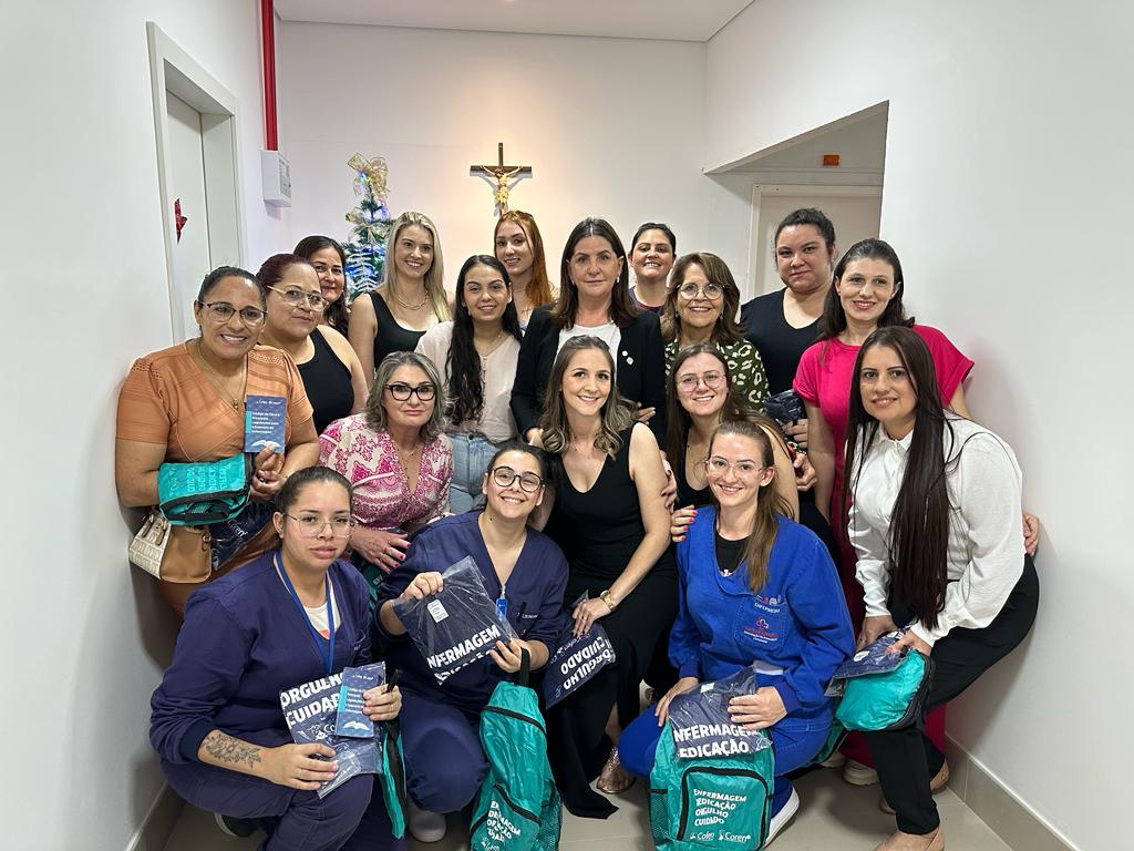Grupo de 18 mulheres reunidas em um corredor de hospital, posando para foto. Algumas usam uniformes azul-marinho ou azul-claro com bordados, enquanto outras vestem roupas casuais ou sociais. Várias seguram mochilas ou sacolas verdes com a inscrição “Enfermagem Educação” e panfletos. Ao fundo, há um crucifixo fixado na parede branca.