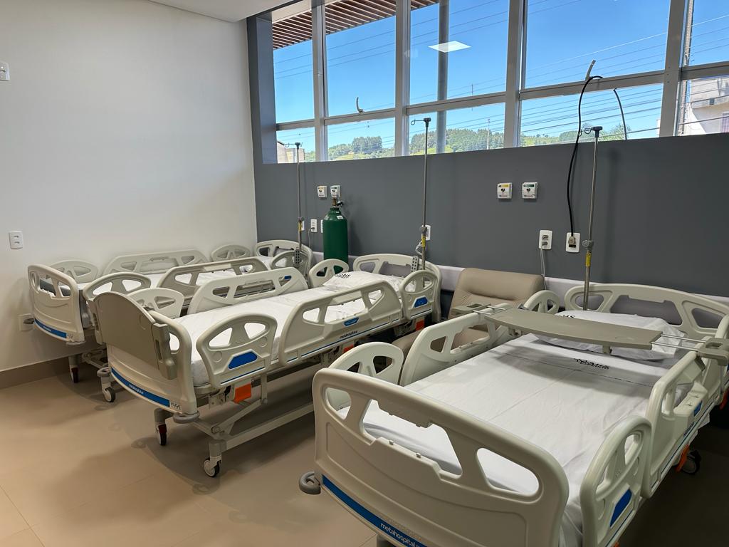 Quarto hospitalar com várias camas articuladas lado a lado, equipadas com grades laterais, travesseiros e lençóis brancos. Há suportes para soro, poltrona de acompanhante e cilindro de oxigênio. A parede cinza possui tomadas e saídas de gases medicinais. Grandes janelas permitem entrada de luz natural.