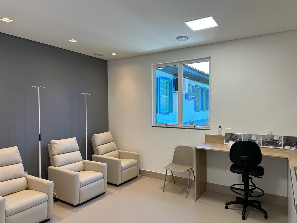 Sala de atendimento hospitalar com três poltronas reclináveis claras alinhadas junto à parede cinza, cada uma acompanhada por suporte de soro. Há uma cadeira plástica e uma mesa de madeira com bancada, sobre a qual estão papéis e um telefone fixo. Uma cadeira de escritório preta está posicionada diante da mesa. A janela ao fundo deixa ver a fachada azul de outro prédio.