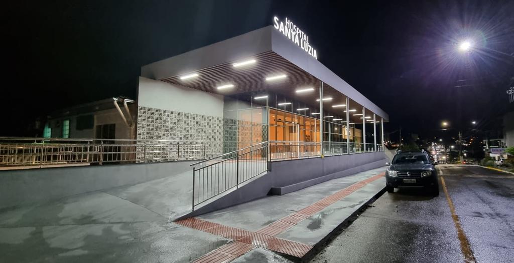 Fachada do Hospital Santa Luzia à noite, com iluminação interna e externa acesa. O prédio possui arquitetura moderna, com grandes janelas de vidro e cobertura com linhas retas. Na frente, há rampa de acessibilidade com corrimãos metálicos e piso tátil. Um carro está estacionado na rua ao lado, e postes iluminam a via.