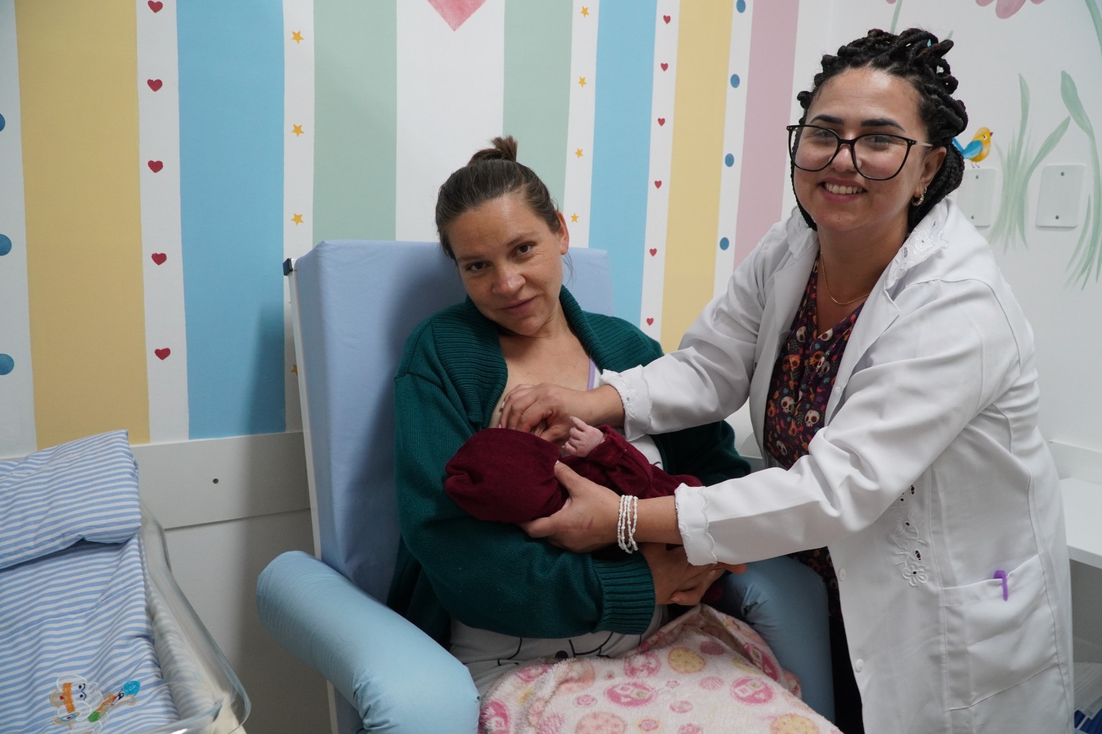 Mãe sentada em cadeira segura o recém-nascido nos braços enquanto profissional de saúde com jaleco branco e óculos auxilia com um sorriso, em ambiente hospitalar infantil decorado com cores e desenhos.