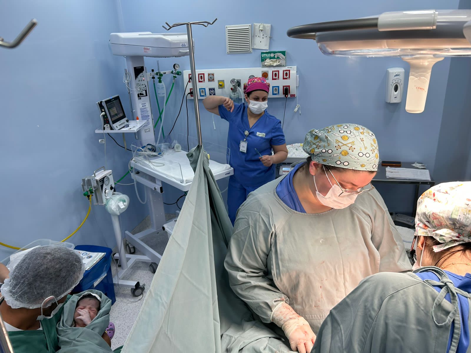 Imagem de uma equipe médica realizando uma cesariana em um centro cirúrgico. À esquerda, um profissional segura o recém-nascido enquanto, ao fundo, há um berço aquecido com equipamentos médicos. Profissionais estão paramentados e o ambiente é esterilizado, com iluminação cirúrgica e paredes azuis.