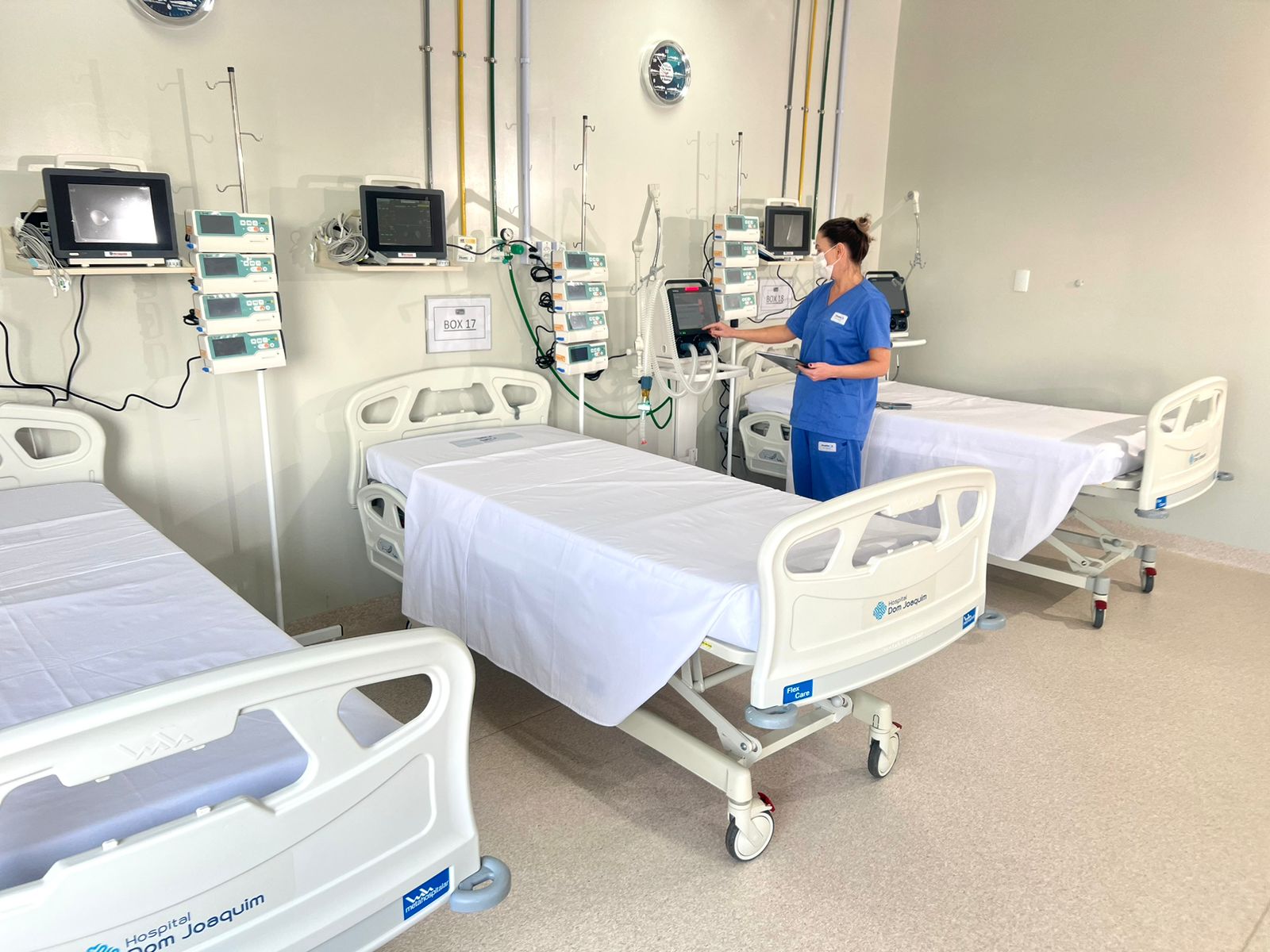 Enfermeira com máscara e uniforme azul mexe em equipamento médico ao lado de um leito hospitalar no Hospital Dom Joaquim. Três camas hospitalares estão alinhadas com lençóis brancos, acompanhadas de monitores e bombas de infusão.