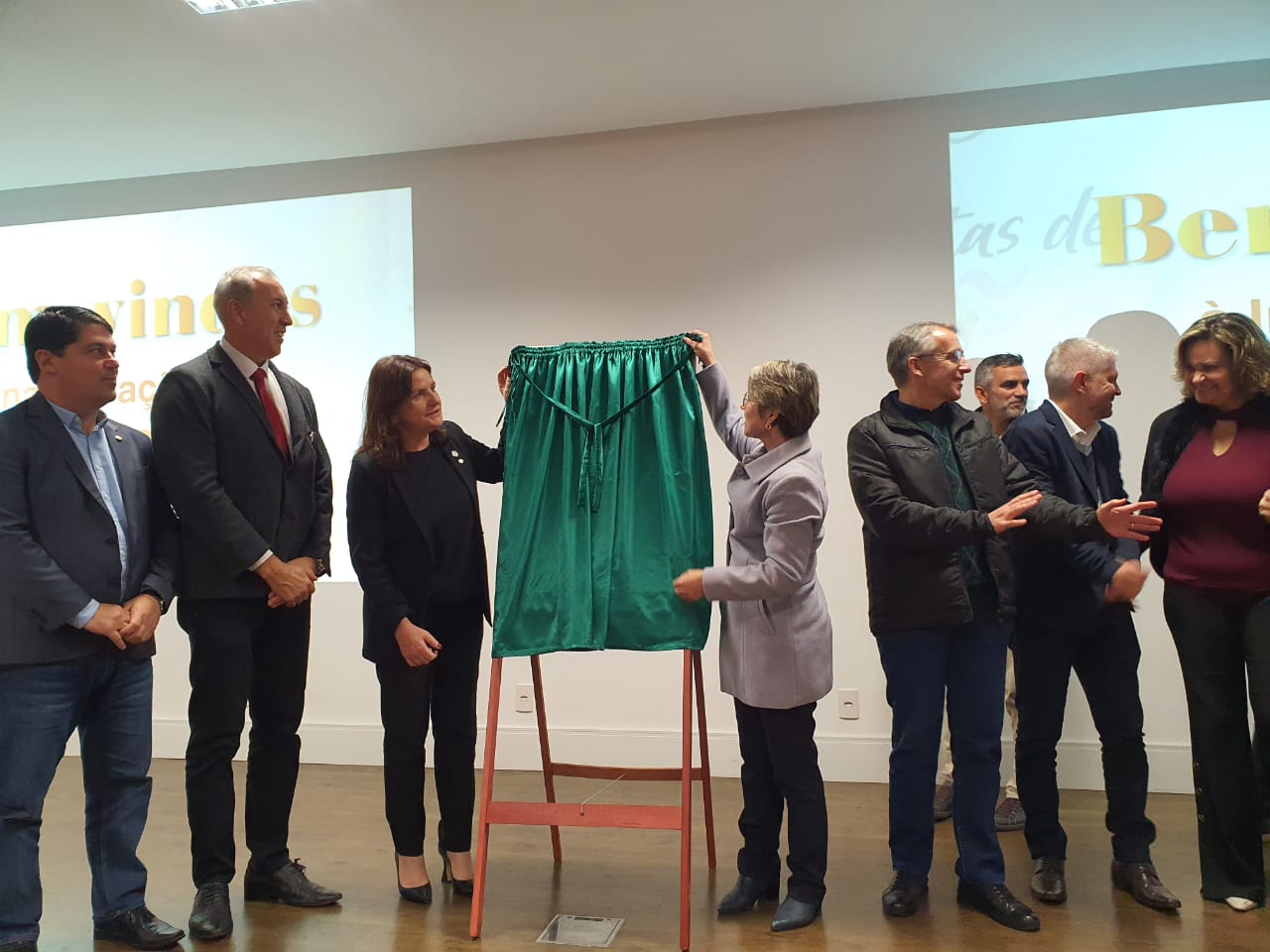 Grupo de autoridades em cerimônia de inauguração ou entrega oficial, reunidos ao redor de um cavalete com uma placa coberta por um pano verde. Duas mulheres seguram o tecido prestes a revelar a placa. O evento ocorre em ambiente fechado, com projetores ao fundo exibindo a palavra 'Bem-vindos'. Todos estão trajando roupas formais.