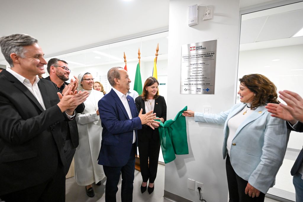 A imagem mostra um grupo de pessoas participando de uma cerimônia de inauguração. No centro, um homem de terno azul e uma mulher de blazer claro seguram juntos um tecido verde, desvelando uma placa prateada fixada na parede. Ao fundo, há outras pessoas sorrindo, incluindo uma religiosa de hábito branco, e duas bandeiras — uma do Brasil e outra de Santa Catarina.  A placa indica a inauguração da Unidade de Internação do Complexo Madre Teresa, em Itajaí (SC), com destaque para autoridades presentes. O ambiente é moderno e bem iluminado, transmitindo um clima formal e comemorativo.