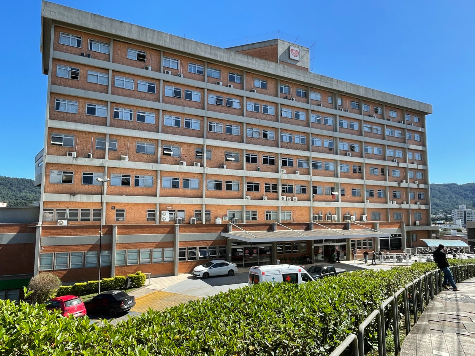 A imagem mostra a fachada principal de um grande hospital de vários andares, construído em tijolos aparentes e concreto, com diversas janelas e aparelhos de ar-condicionado instalados.  Na parte inferior, há uma entrada coberta, por onde passam ambulâncias e carros de passeio, com uma ambulância estacionada no momento. Em frente à entrada, há um jardim com arbustos bem cuidados e uma calçada com corrimão. Ao fundo, vê-se um céu azul e áreas verdes, sugerindo que o hospital está localizado em uma região com relevo e vegetação.
