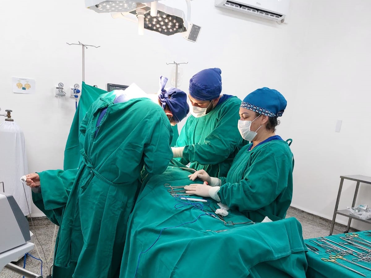 Equipe médica realizando cirurgia em centro cirúrgico, com três profissionais usando avental e touca cirúrgica, máscaras e luvas, manipulando instrumentos sobre paciente coberto por campo estéril.