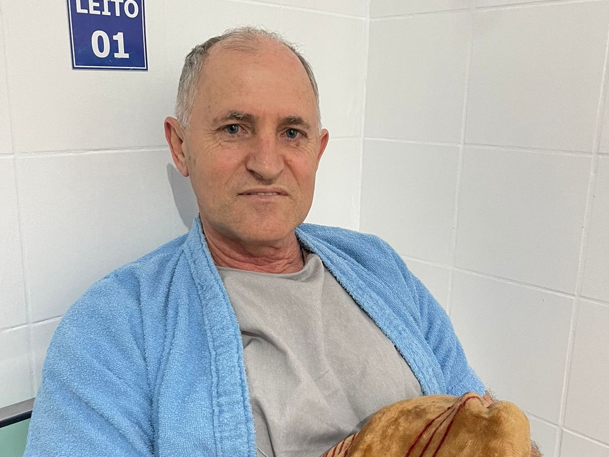Homem de meia-idade sentado em leito hospitalar, vestindo roupão azul claro sobre camiseta cinza, segurando cobertor bege. Ao fundo, parede branca com placa azul indicando "LEITO 01".