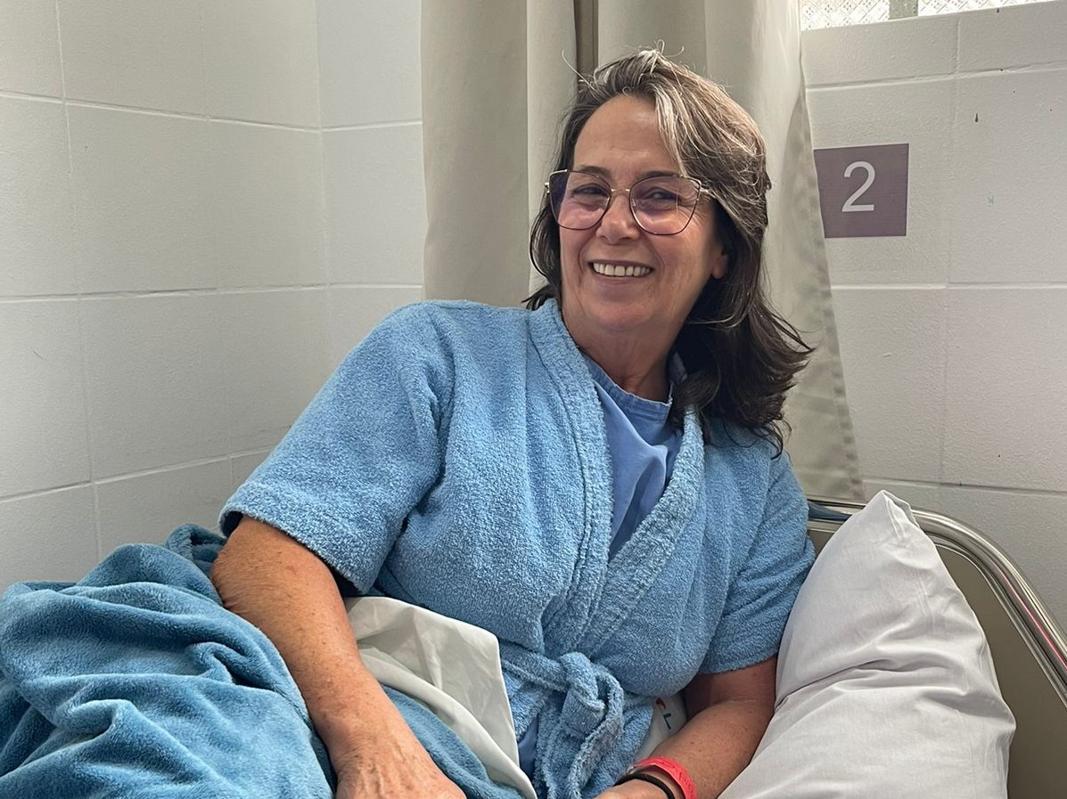 Mulher de meia-idade, sorridente, deitada em leito hospitalar, vestindo roupão azul claro e cobertor azul. Ao fundo, cortina clara e número 2 na parede indicando o leito. Ambiente limpo e iluminado.