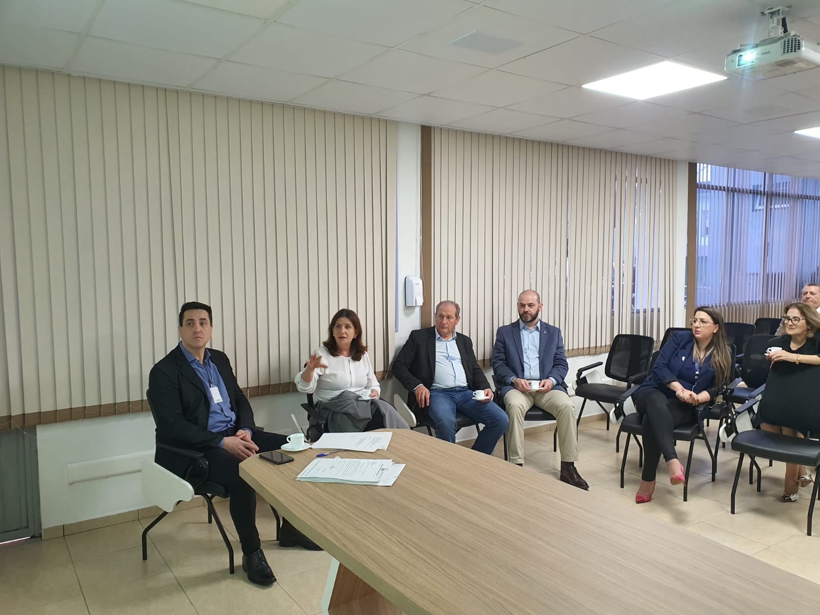 Homens e mulheres participam de uma reunião em uma sala com persianas fechadas. Sentados em semicírculo, alguns seguram xícaras de café enquanto ouvem atentamente uma mulher que fala com gestos. Papéis e celular estão sobre a mesa ao centro.