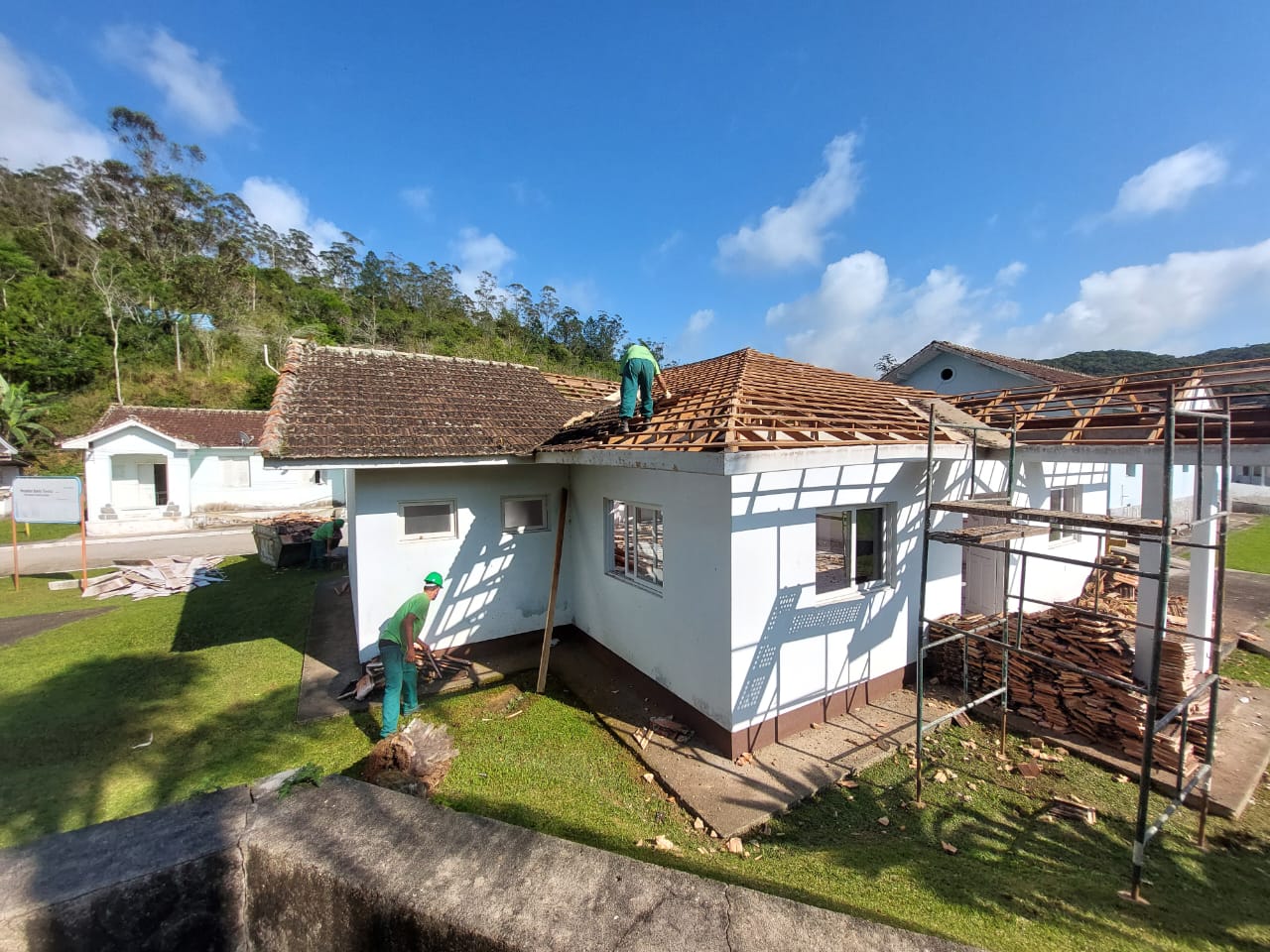 Trabalhadores removendo parte do telhado de um prédio branco com telhas marrons. Um operário está no telhado, enquanto outro recolhe materiais no chão. Há andaimes montados ao lado da estrutura e um gramado ao redor. Ao fundo, árvores e céu azul com nuvens.