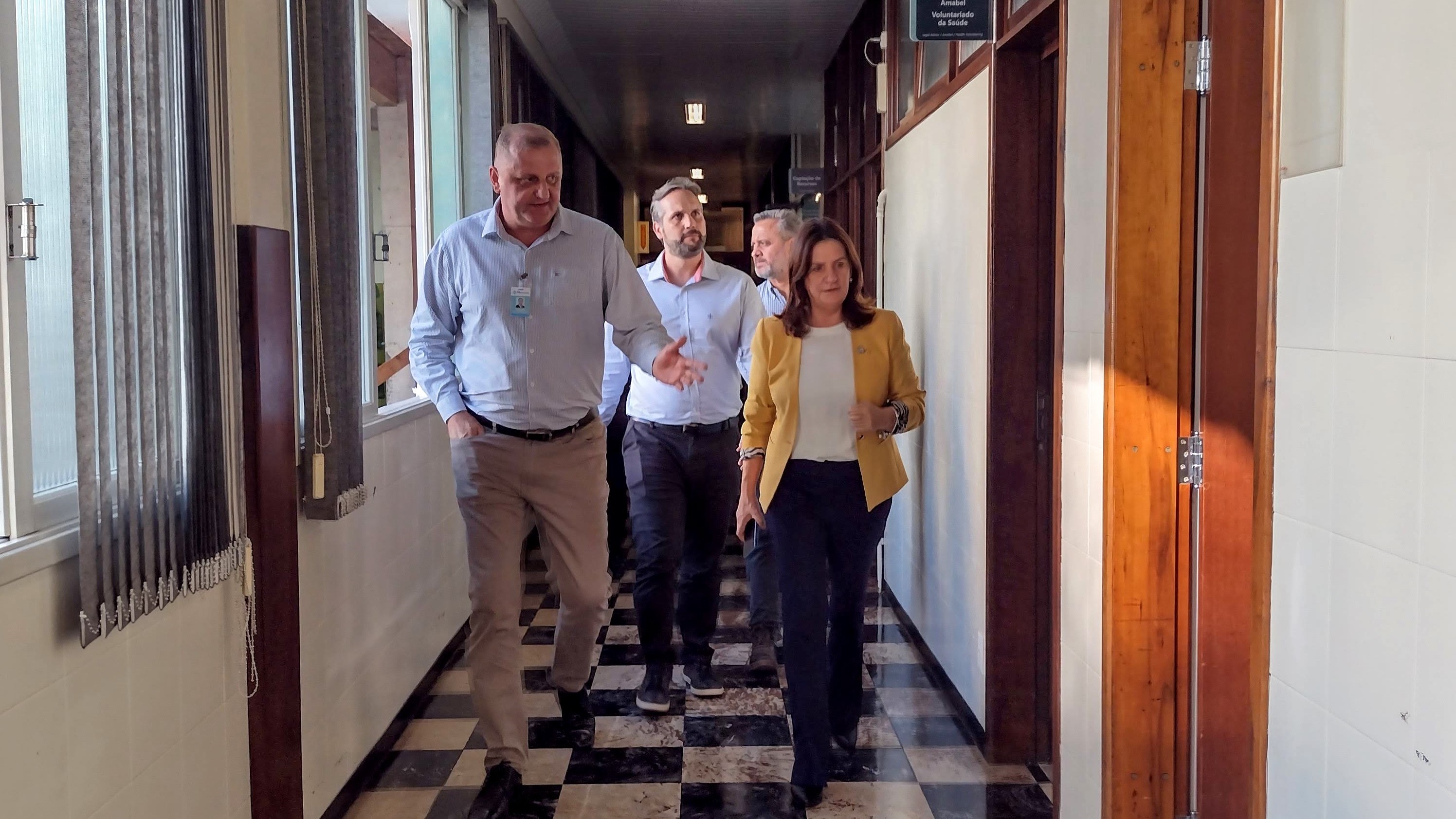 Grupo de pessoas caminhando por um corredor interno com piso quadriculado em preto e branco. À frente, um homem conversa com uma mulher de blazer amarelo, acompanhados por outros homens em trajes sociais. O ambiente é bem iluminado por janelas laterais.