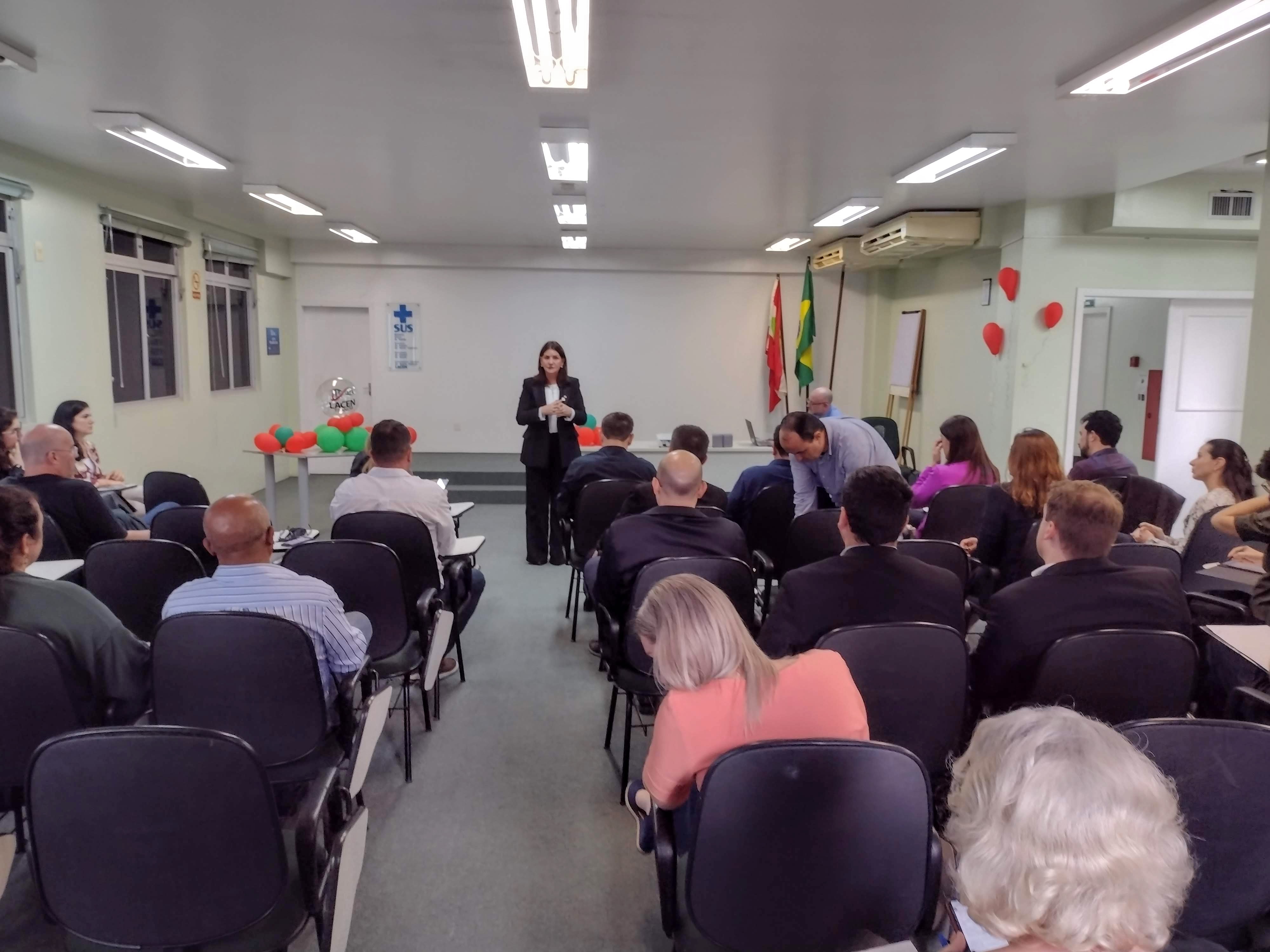 Sala de reuniões com cadeiras organizadas em fileiras. Servidores assistem a uma mulher em pé, à frente, falando durante o evento. No fundo, há balões nas cores verde e vermelho e bandeiras do Brasil e de Santa Catarina.