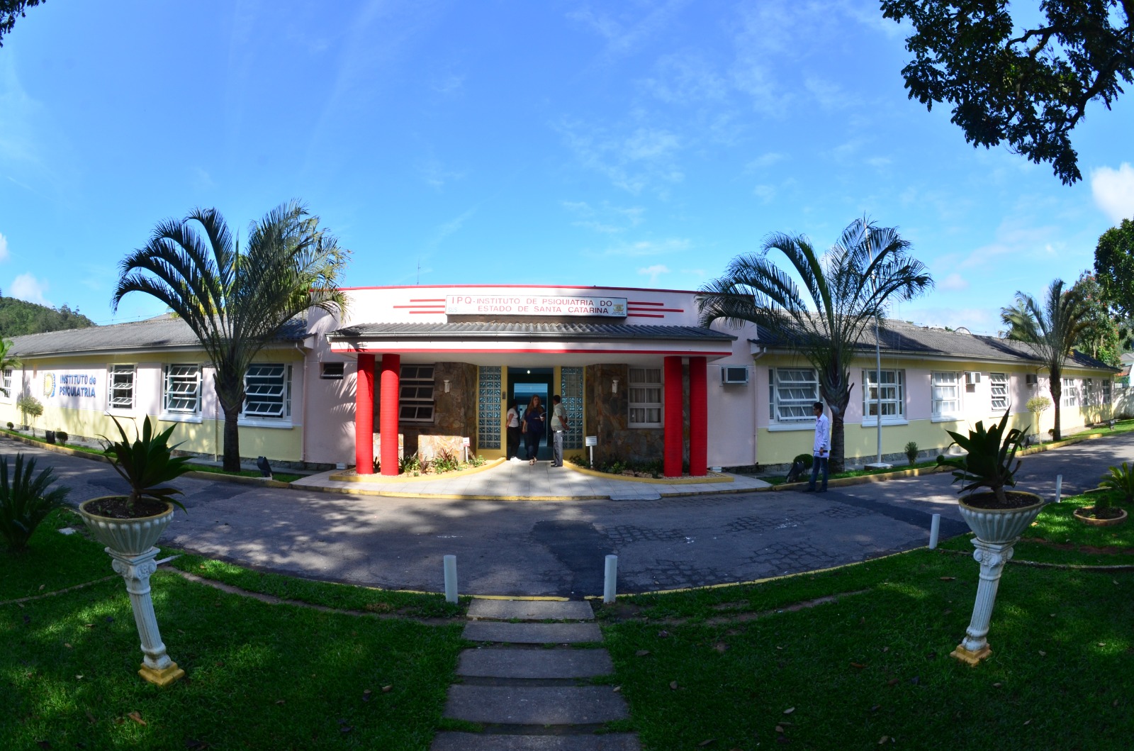 A imagem mostra a fachada do Instituto de Psiquiatria do Estado de Santa Catarina (IPQ), um prédio térreo de arquitetura simples, com destaque para a entrada central marcada por duas colunas vermelhas e um letreiro com o nome da instituição. O local está cercado por um jardim bem cuidado, com palmeiras e vasos ornamentais, transmitindo uma aparência acolhedora. O céu azul e o clima ensolarado ajudam a dar um aspecto convidativo e tranquilo à cena.