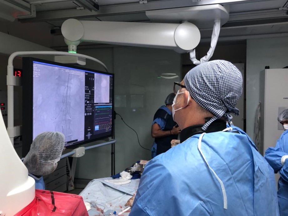 Equipe médica em centro cirúrgico realiza procedimento com auxílio de equipamento de imagem. Profissional em primeiro plano observa monitor com exames em tempo real. Todos estão usando roupas cirúrgicas, toucas e máscaras, indicando ambiente estéril e de alta tecnologia.